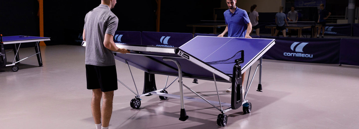 Two people setting up a portable ping pong table in an indoor setting with a visible brand logo.