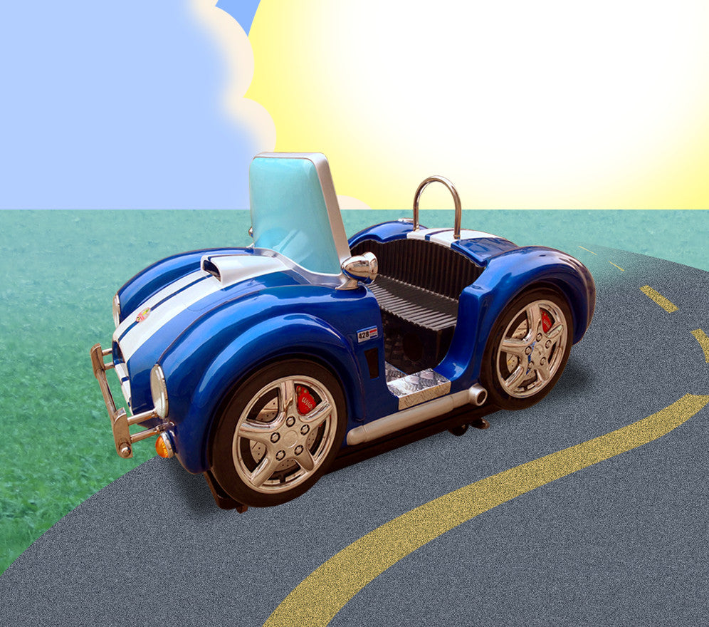 A blue and white toy model of a KC Cobra classic car positioned on a road with a grassy background and a sunny sky overhead.