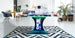 An ICE Super Chexx Bubble Hockey table with a blue base and red grips, featuring custom 40th graphics and LED lighting, placed in a modern room with plants and furniture around it.