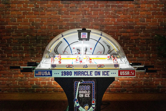 An illuminated dome hockey table featuring custom graphics for Team USA and Team Canada, with a transparent dome and a red base.