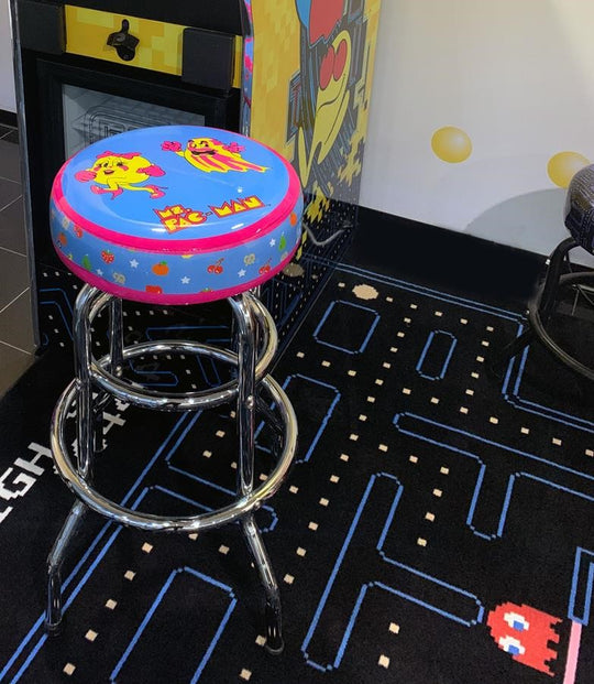 A colorful bar stool with a Pac-Man design, featuring characters and motifs from the game, placed in a game room setting with an arcade machine in the background.