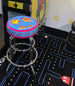 A colorful bar stool with a Pac-Man design, featuring characters and motifs from the game, placed in a game room setting with an arcade machine in the background.