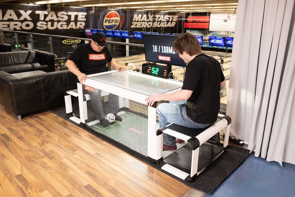 Two people playing air hockey in an indoor setting with various advertisements on the walls.